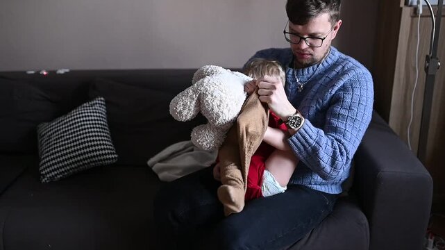 Young father dressing his toddler child while sitting on a sofa at home. Everyday family life moment, parenting routine, care and bonding. Child slightly resists while getting dressed, realistic lifes
