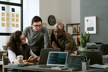 Multiethnic group of young adult men and man collaborating around laptops in modern office, discussing project while analyzing code on computer screens, working together on software development