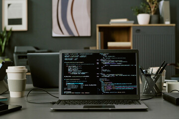 Laptop displaying programming code on screen positioned on office desk with takeaway coffee cups,...