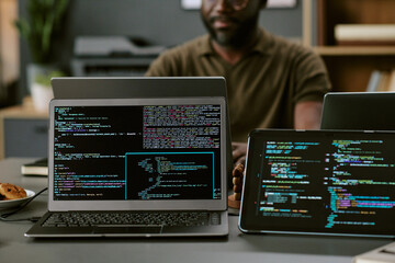 Black man sitting at desk working on laptop with multiple screens displaying programming code, focusing on software development in modern office environment, partial face visible
