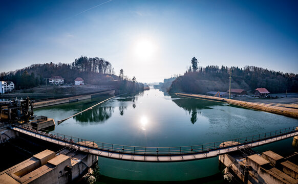 Aerial view of a tranquil river reflecting the sun, framed by verdant hills and a concrete dam with a pedestrian bridge, Hagneck, Bern, Switzerland.