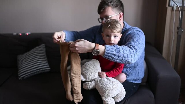 Young father dressing his toddler child while sitting on a sofa at home. Everyday family life moment, parenting routine, care and bonding. Child slightly resists while getting dressed, realistic lifes