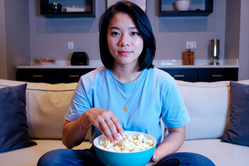 Portrait of young adult Asian woman sitting on sofa eating popcorn while watching television, holding bowl in lap, looking directly at camera in modern living room setting