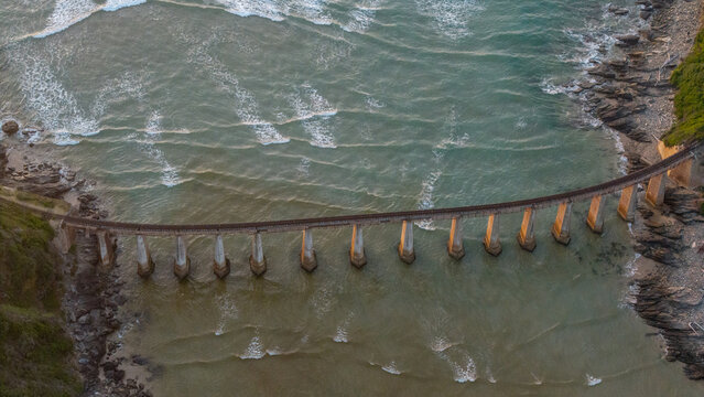 Aerial view of a bridge stretching over the ocean, contrasting with the rugged coastline and the soft hues of the water, Wilderness, Western Cape, South Africa.