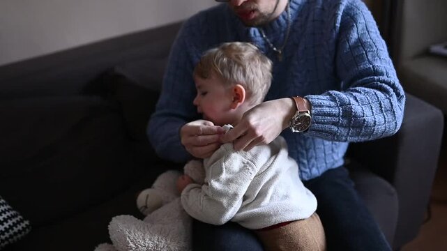 Young father dressing his toddler child while sitting on a sofa at home. Everyday family life moment, parenting routine, care and bonding. Child slightly resists while getting dressed, realistic lifes