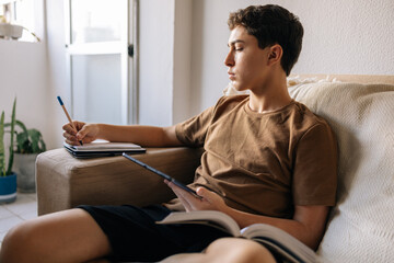 Teenage boy sitting on a sofa at home, reading a book, studying and writing in a notebook