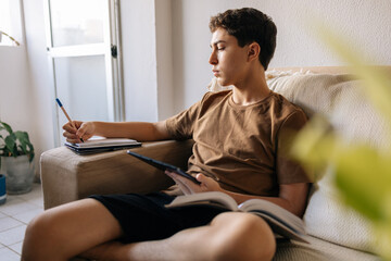 Teenage boy sitting on a sofa at home, reading a book, studying and writing in a notebook