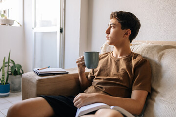 Teenage boy sitting on a sofa at home holding a cup of hot drink. Cozy lifestyle, comfort, rest, study break and quiet moment indoors