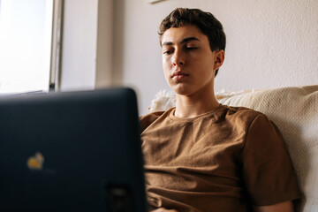 Teenage boy using a laptop while sitting on a sofa at home. Remote study, technology, digital life and modern lifestyle concept