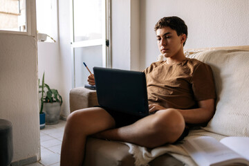 Teenage boy using a laptop while sitting on a sofa at home. Remote study, technology, digital life and modern lifestyle concept
