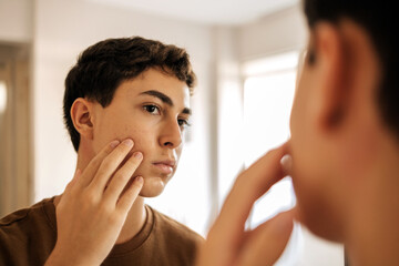 Teenage boy looking at himself in the mirror and applying skin care product on his face. Acne, self care, hygiene and adolescent routine concept