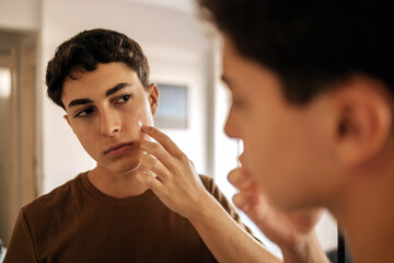 Teenage boy looking at himself in the mirror and applying skin care product on his face. Acne, self care, hygiene and adolescent routine concept