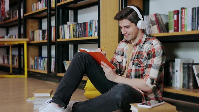 Young smart intelligent college student boy wearing casual clothes listen to music in headphones write in notebook study visit university library or book store sitting near shelf. Knowledge concept