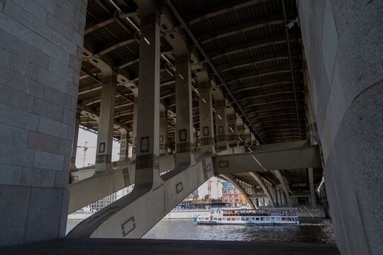 View from under the Andreevsky bridge showcasing its concrete pillars and metal framework. A white riverboat with blue trim moves along the river beneath the bridge. City buildings and construction cr