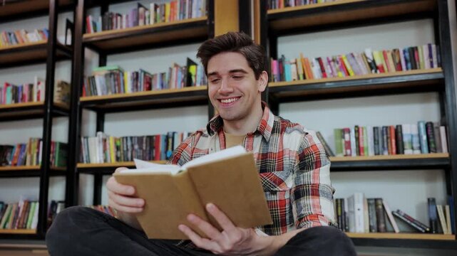 Young smiling cheerful intelligent college student boy wears casual clothes studying visit university library sitting on floor near shelf reading interesting book prepare for exam. Knowledge concept