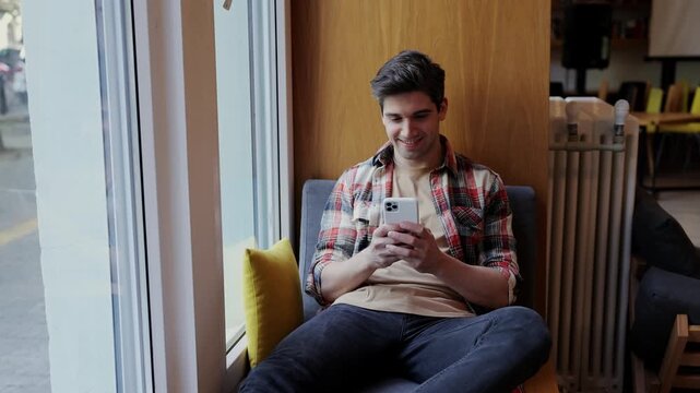 Young happy smart intelligent college student boy wears casual clothes studying visit university library sitting in chair hold in hand use mobile cell phone search new information. Knowledge concept