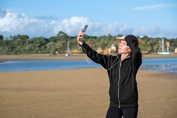 Sporty woman in a black hoodie taking a selfie with a smartphone while wearing white earbuds