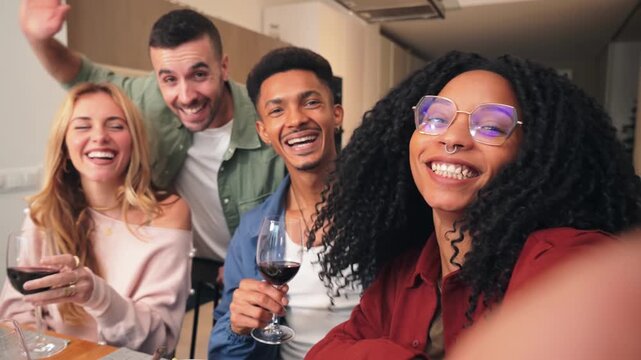A cheerful group of friends happily celebrating together, sharing laughter and joy while taking a moment to wave and look at the camera during their delightful gathering with wine glasses raised.