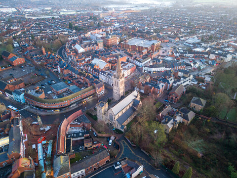 Aerial view of St Andrew's Church spire piercing through the urban sprawl, brick buildings contrasting with green spaces under a soft, diffused light, Rugby, England, United Kingdom.