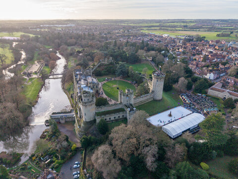 Aerial view of the majestic Warwick Castle, its stone walls softened by the surrounding emerald foliage, standing proud against the tranquil River Avon, Warwick, England, United Kingdom.
