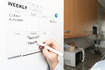 Woman writing household tasks on a weekly planner attached to the kitchen refrigerator