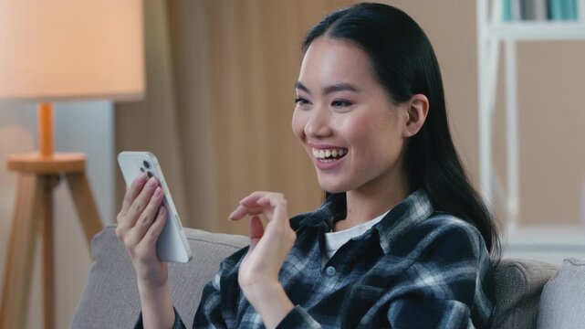 Smiling Asian woman holding smartphone at home happy customer female girl using mobile payment app making online purchase excited lady nodding Korean Japanese Chinese shopper browsing cellphone indoor