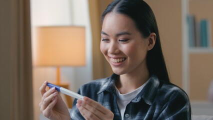 Happy pregnant Asian Korean woman surprised holding positive pregnancy test at home expectant mother smiling female girl looking result childbirth parenthood healthcare future mom baby infant family