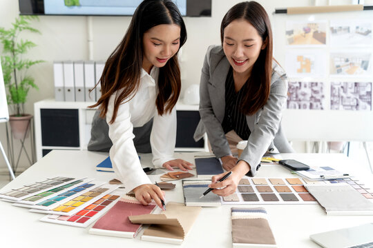 Two Asian adult designers collaborate on material selection choosing fabric samples color palettes for interior project in bright office environment