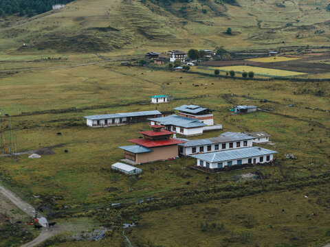 Aerial view of buildings nestled in the vast, golden-green valley under a soft sky, with mountains rising gently in the distance, Phobjikha Valley, Wangdue Phodrang, Bhutan.