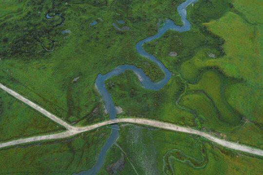 Aerial view of a meandering river cutting through a lush green valley floor, intersected by a narrow path, Phobjikha Valley, Wangdue Phodrang, Bhutan.