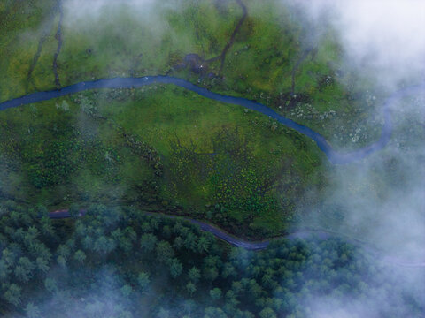 Aerial view of a winding river cutting through the lush green valley, embraced by ethereal clouds and dense forest, Phobjikha Valley, Wangdue Phodrang, Bhutan.