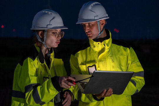 Workers discuss project details during night shift at construction site with laptop and tools in hand