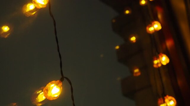 Festive Christmas Garland Features Dazzling Orange Lights In Shape Of Small Pumpkins, Casting A Warm Glow Through Window, Enriching Night Sky With Whimsical Cheer And Holiday Spirit. Christmas Xmas