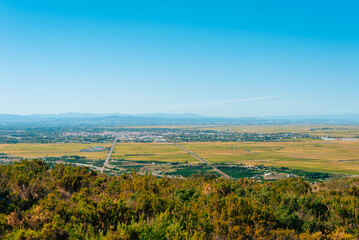 A wide view shows a city surrounded by fields and distant mountains. The scene captures a clear day with blue sky. Roads line the fields, connecting the landscape
