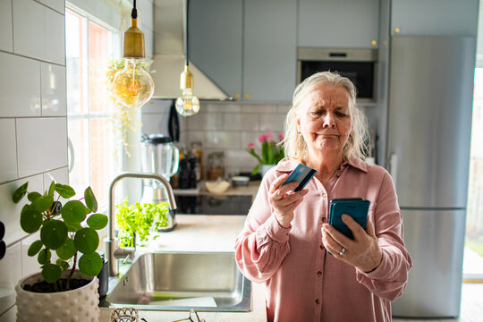 Senior woman using credit card on smartphone in home kitchen