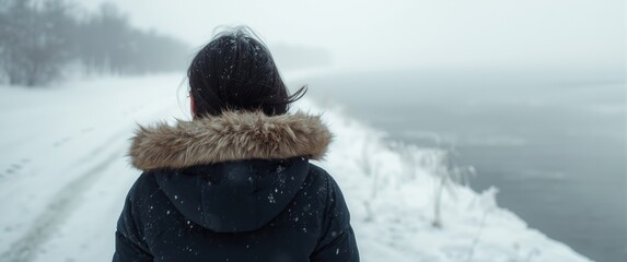 Back view of woman wearing a winter jacket and hood strolling on frozen riverbank during a windy, sunny winter day with a blizzard