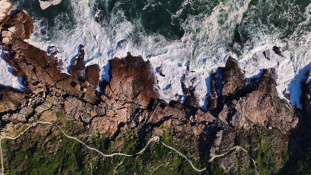 Aerial view of the rugged coastline where the foamy white waves crash against the dark rocks, and a walking path meanders along the lush greenery, Cape Town, Western Cape, South Africa.