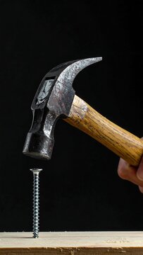 Close-up view of a hand using a claw hammer to drive a screw into a piece of wood against a black background