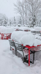 Outdoor cafe tables and chairs buried under thick, fresh snow during a heavy winter blizzard, indicating extreme cold weather and a strong winter storm