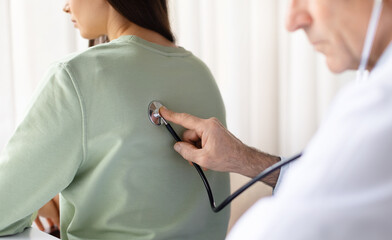 Male doctor performing back examination with stethoscope on female patient during medical checkup, therapist man listening breathing of young woman during appointment in clinic