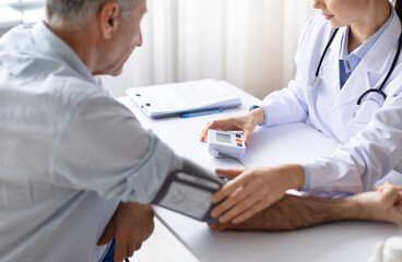 Female doctor measuring blood pressure of senior male patient using digital monitor during routine medical checkup in clinic setting, closeup shot