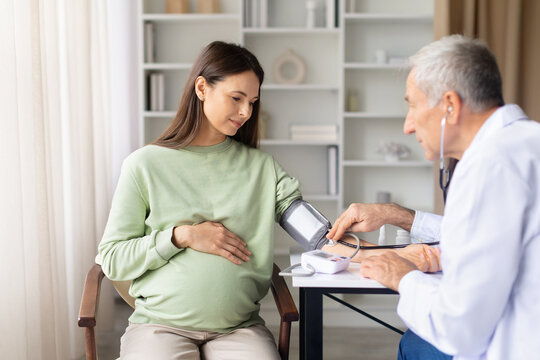 Senior male doctor measuring blood pressure of pregnant woman during prenatal examination, maternity health monitoring and routine pregnancy care