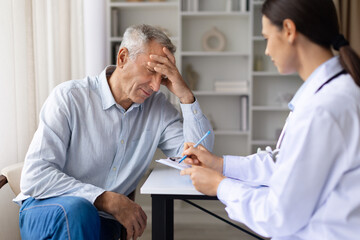 Senior male patient experiencing headache while female doctor records symptoms during medical consultation in clinic office environment, writing notes to clipboard