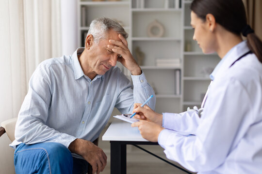 Senior male patient experiencing headache while female doctor records symptoms during medical consultation in clinic office environment, writing notes to clipboard