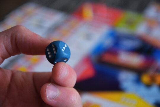 Close up of hand holding a blue d6 dice over a blurred board game