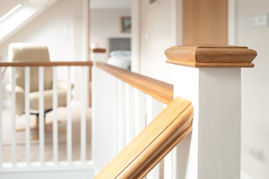 Shallow focus of a first floor wooden bannister seen on the top floor of a recent loft conversion. A recliner chair is on the landing and a bedroom in the distance.