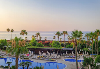 Serene tropical resort view at sunset. A large, blue swimming pool in the foreground, surrounded by loungers and palm trees, with the calm ocean stretching to the horizon under a soft, pastel sky