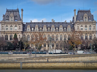 Paris, France - December 10, 2025: Beautiful view of the Hotel de Ville from the Seine riverside, showing the grand Neo-Renaissance facade and the riverbanks lined with trees and stone embankments
