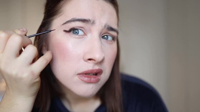 Young woman practices drawing eyeliner but nothing turns out right looking worried and discouraged as she studies her uneven line up close trying to understand what went wrong
