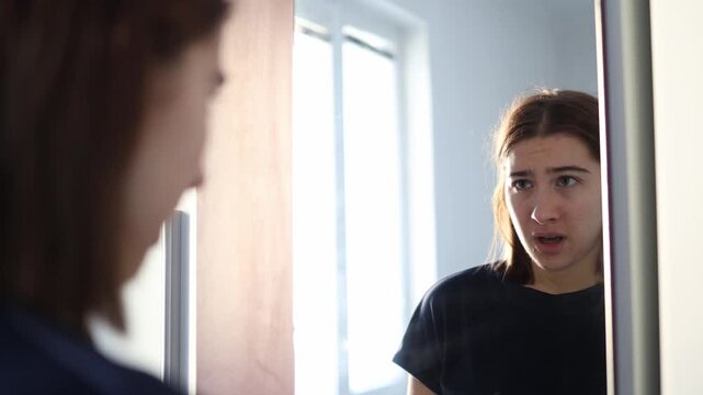 Young woman stands at the mirror feeling insecure as she examines her face imagining many imperfections reacting with doubt and vulnerability in soft morning light inside her quiet room